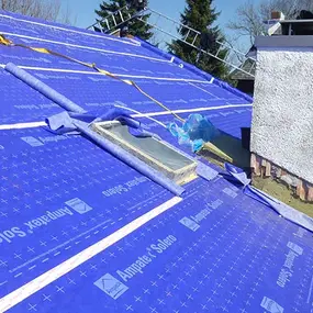 Dachsanierung mit blauer Unterspannbahn, Dachfenster und Schornstein bei sonnigem Wetter.