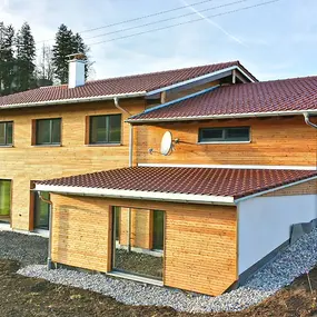 Modernes Einfamilienhaus mit Holzfassade, roten Dachziegeln und großen Fenstern im Grünen.